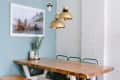 Dining area with a wooden table, gold pendant lights, framed beach photo, and eucalyptus in a vase.