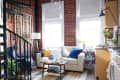 Loft-style living room with exposed brick, cream sofa, blue and yellow pillows, spiral staircase, and modern decor.