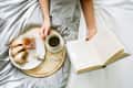 Person reading a book in bed with a tray of coffee, bread, and jam.