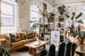 Loft living room with yellow sofa, geometric pillows, wooden coffee table, and plants on shelves against brick walls.