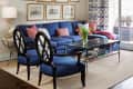 Living room with blue sofa, pink pillows, glass coffee table, framed art, and patterned curtains near a large window.
