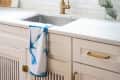 White kitchen with gold faucet, blue-trimmed window, and blue tie-dye towel on sink.