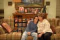 Two people sitting on a plaid sofa with a colorful crochet blanket, surrounded by family photos and a dinosaur figurine.