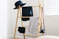 Wooden clothes rack with a black hat, dark and light shirts, and a camera strap in a bright room.