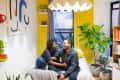 Couple sitting on a bed in a bright room with yellow walls, plants, bookshelves, and abstract wall art.