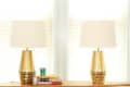 Two gold table lamps with white shades on a wooden table, next to stacked books and a small plant.