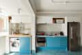 Modern kitchen with blue cabinets, stainless steel appliances, red kettle, and potted plants on wooden shelves.