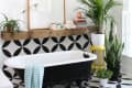 Clawfoot bathtub with black and white geometric tiles, wooden shelf, potted plants, and a striped towel.