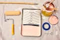 Paint supplies on a drop cloth, including a roller, tray with pink paint, brush, tape, and open paint can.