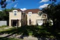 Two-story beige house with brown roof, large front lawn, and a real estate sign.