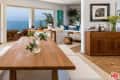 Open-plan living and dining area with ocean view, wooden table, white benches, potted plants, and modern decor.
