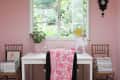 Home office with pink walls, white desk, floral chair cover, and window view. Two wooden chairs with stacked books.