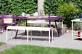 Purple outdoor dining table with white benches, set with dishes and drinks, surrounded by greenery and a basket of apples.