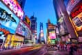 Times Square at dusk with vibrant billboards, bustling crowds, and blurred lights from passing traffic.