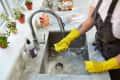 Person wearing yellow gloves cleaning a kitchen sink with a sponge, surrounded by potted plants and cleaning supplies.