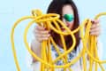 Person in white dress holding tangled yellow extension cord against blue wall.