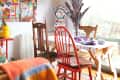 Eclectic dining area with colorful chairs, a patchwork table, vibrant wall art, and a mannequin in a bright room.