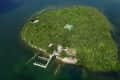 Aerial view of a lush green island with a dock, sandy beach, and tennis court surrounded by water.