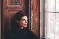 Woman in black top sitting by a wooden window, looking outside thoughtfully.