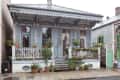Historic blue house with ornate trim, shuttered windows, and a porch adorned with potted plants and flowers.