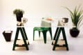 Glass desk with green sawhorse legs, a small plant, mug labeled "Ann," sandwich, clock, and green chair.