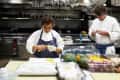 Chefs in a commercial kitchen preparing vegetables, with hanging pots and pans in the background.