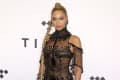Woman in a sheer black beaded gown with a long braided ponytail, standing against a backdrop with geometric patterns.