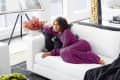 Woman in purple outfit lounging on a white sofa in a modern living room with large windows and a table lamp.