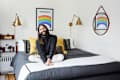 Person sitting on a bed with yellow and black pillows, rainbow artwork, and brass wall lamps in a cozy bedroom.
