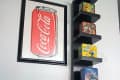 Framed Coca-Cola can art above a vertical shelf with vintage lunchboxes, next to a metal pet bowl on wooden floor.