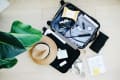 Open suitcase with camera, striped shirt, hat, sandals, and toiletries on a wooden floor next to a large green plant.