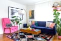 Living room with a navy sofa, pink armchair, colorful rug, and plants, featuring framed art and a wooden coffee table.