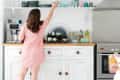 Woman in a pink dress reaching for a mug on a kitchen shelf above a white cabinet with various dishes and decor.