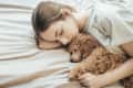 Woman sleeping on a bed, cuddling a small brown poodle.