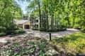 Two-story house with stone facade, surrounded by lush trees and blooming pink flowers, featuring a curved driveway.