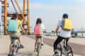 Three people cycling on a waterfront path, each wearing colorful backpacks: teal, pink, and yellow.