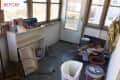 Cluttered sunroom with white shelf, paint cans, ceiling fan parts, and potted plant near a door and windows.