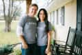 Couple in matching gray "#shiplap" shirts standing on a porch with black rocking chairs.