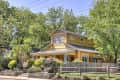 Yellow two-story house with a wraparound porch, surrounded by lush green trees and a wooden fence.