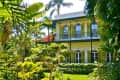 Two-story yellow house with green shutters, surrounded by lush tropical plants and palm trees.