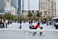 People ice skating at an outdoor rink in a city park, surrounded by tall buildings and palm trees.