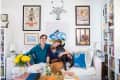 Couple sitting on a white sofa with a black dog, surrounded by colorful art, bookshelves, and yellow flowers in a vase.