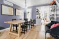 Dining room with wooden table, black chairs, purple walls, and framed art, leading to a bright kitchen with plants.