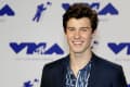 Young man in a dark suit and patterned shirt smiling at the 2017 VMA Awards backdrop.
