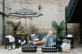 Outdoor patio with black and white furniture, striped umbrella, plants, and a woman sitting on a patterned ottoman.