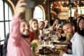 Group of people at a restaurant table, smiling and taking a selfie, with wine glasses and plates of food.