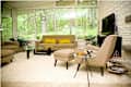 Mid-century modern living room with beige sofa, armchairs, glass coffee table, and large windows overlooking greenery.