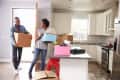 Couple moving boxes into a modern kitchen with white cabinets and stainless steel appliances.