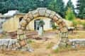 Stone archway leading to a rustic house with a circular window, surrounded by trees and greenery.