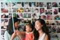 Two women with a toddler in a coral dress, holding balloons, in front of a photo collage wall.